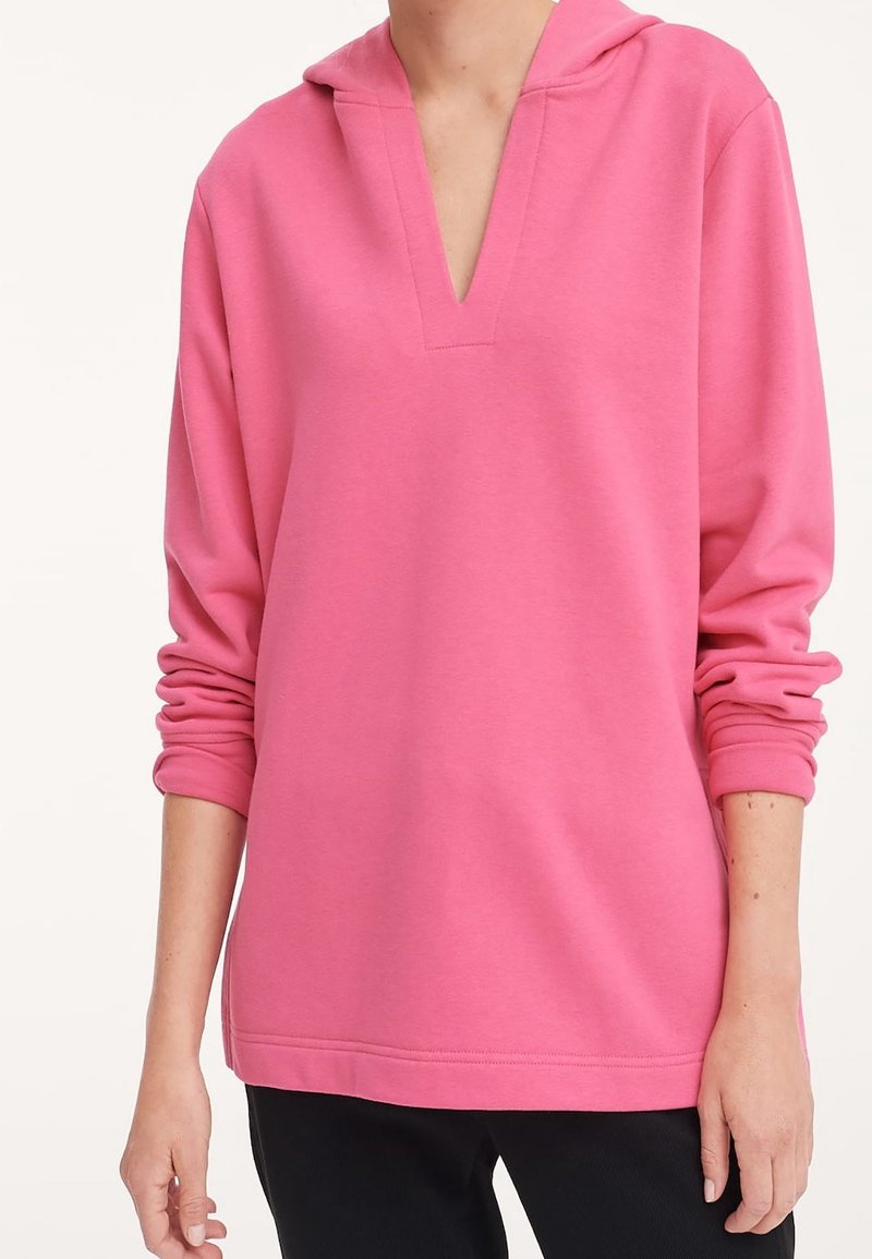 Personne portant un pull rose ample à manches longues avec un col en V profond et une capuche, associé à un pantalon noir.