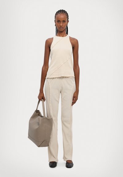 Beige ribbed tank top with diagonal seam details, paired with light beige pants featuring side stitching and raw edges, holding a grey tote bag.