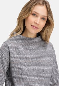 Haut à carreaux gris avec un col montant, tissu texturé et coupe ample. Présente un subtil accent marron dans le motif à carreaux.