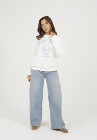 Donna in piedi con una felpa bianca "New York", jeans azzurri a gamba larga e scarpe sportive grigie su uno sfondo chiaro e semplice.