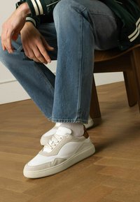 Witte en beige sneakers met suede accenten en een gestructureerde zool, gecombineerd met blauwe denim en witte sokken, zittend op een houten vloer.