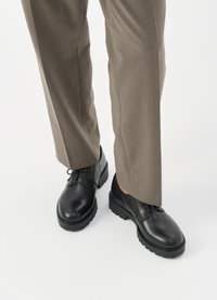 Scarpe di cuoio nere con una finitura liscia, dotate di una suola spessa e design con lacci, abbinate a pantaloni su misura color marrone chiaro.