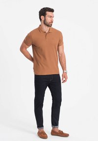 Hnědá polokošile s límečkem a vyšitým logem, v kombinaci s tmavými džíny a hnědými mokasínami. Model má krátké vlasy a strniště.