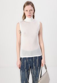 Hvit ermeløs turtleneck topp laget av gjennomsiktig stoff, sammen med mørk denim bukser med metallglidelåsdetaljer. Lys grå håndveske i hånden.