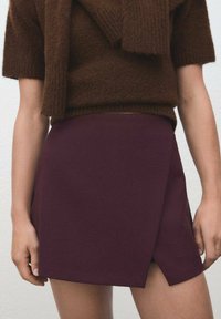Donna che indossa un maglione lavorato a maglia marrone con una sciarpa coordinata e una gonna alta di vita di un viola scuro con uno spacco frontale, in piedi su uno sfondo neutro.