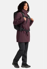 Outdoor jacket in dark purple, made of soft fabric, featuring a front zipper, hood, and adjustable waist. Model wears black backpack and pants.