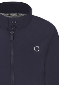 Giacca blu navy con colletto alto, dotata di zip frontale, due bottoni e un logo circolare sul petto. L'interno ha una fodera con motivo.