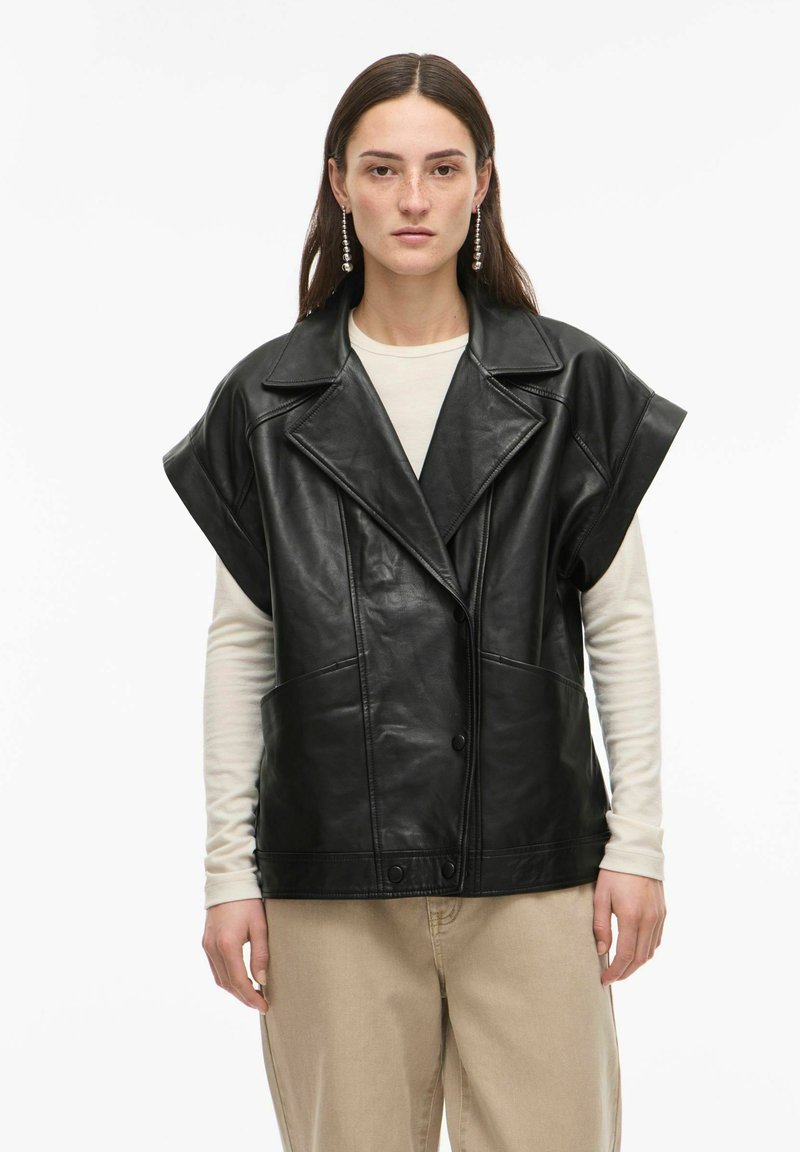 Veste oversize noire en cuir avec manches larges, col à revers pointu et deux poches avant. Portée sur une chemise à manches longues couleur crème, associée à un pantalon clair.