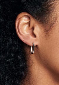 Srebrna kolczyk w kształcie wydłużonego owalu, błyszczące wykończenie, z gładką strukturą. Noszony na uchu modelki z widocznymi ciemnymi kręconymi włosami.