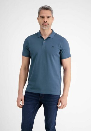 Man met grijs haar, gekleed in een teal korte mouwen poloshirt en donkerblauwe spijkerbroek, tegen een effen witte achtergrond.