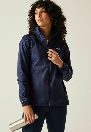 Mujer con cabello rizado que lleva una chaqueta impermeable azul marino, sosteniendo una botella de agua plateada y mirando hacia un lado contra un fondo liso.