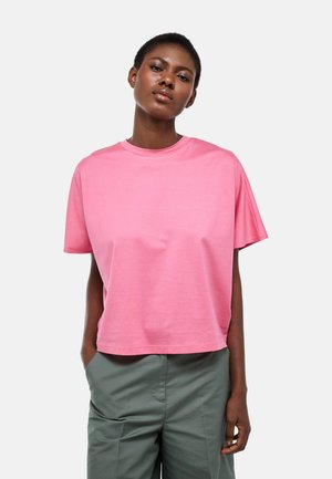 Femme aux cheveux courts portant une chemise rose à manches courtes ample et un pantalon vert, debout les mains dans les poches devant un fond uni.