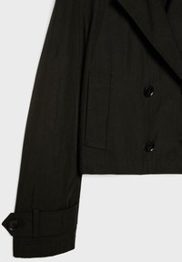 Manche d'une veste noire courte avec poignet boutonné et poche à rabat sur le devant, doublée croisée avec deux boutons visibles sur le torse.