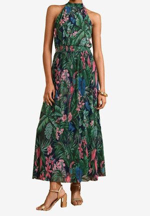 Femme portant une robe sans manches, col haut, longue jusqu'aux chevilles, avec un motif floral tropical vert et des sandales à talons blocs dorés.
