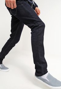 Jeans de mezclilla azul oscuro con costuras en contraste, ajuste slim y textura descolorida. Combinados con zapatos gris de tipo slip-on y calcetines blancos.