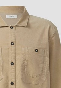 Béžová košová bunda vyrobená z texturovanej látky, s golierom, dvoma náprsními vreckami a čiernymi patentkami. Značka na golieri.