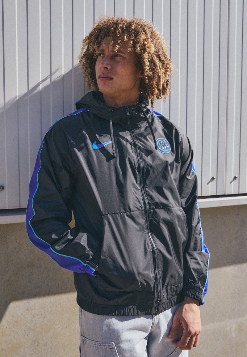 Ung mand med krøllet hår iført en sort og blå Nike-jakke, der står udendørs mod en lys væg og kigger til siden.