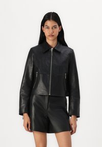 Svart cropped jakke i teksturert lær med en glatt overflate, med glidelås foran og to glidelåslommer, parret med svarte shorts.