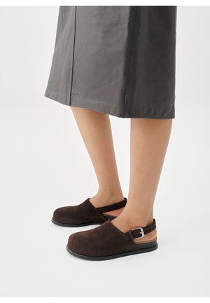 Chaussures slingback en daim marron à bout fermé avec détail de boucle, portées avec une jupe grise longueur genou, présentées sur fond blanc.