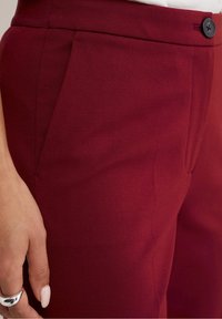 Burgundy byxor med en slät textur, med snedställda fickor och en svart knappknäppning vid midjan. Tyget verkar hållbart och välsittande.