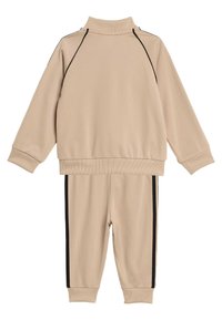 Bež trenirka iz mehke tkanine, ki vključuje visok ovratnik puloverja s črnimi stranskimi črtami na hlačah. Rebrasti manšeti in pas.