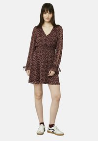 Robe portefeuille florale bordeaux avec de longues manches transparentes, taille froncée et ourlet à volants. Portée avec des baskets blanches et des chaussettes bordeaux.