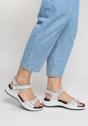 Feet wearing light blue jeans and silver open-toe sandals with adjustable straps and black cushioned soles against a light background.