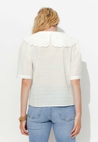 Blouse en coton blanche avec un motif de broderie anglaise, col festonné, manches courtes et ourlet droit, associée à un jean bleu clair.