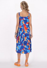 Ärmelloses blaues Kleid mit buntem abstraktem Muster, gestuftem Design und gerafftem Rücken. Kombiniert mit orangefarbenen Riemchensandalen.