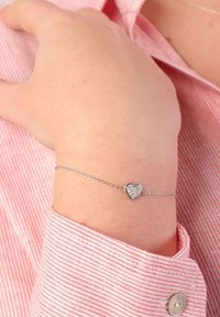 Zilveren schakelarmband met een hartvormige hanger met ingesloten heldere edelstenen, gedragen om een pols, gecombineerd met een gestreept roze shirt.