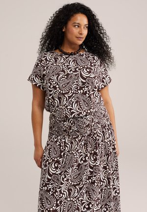 Femme aux cheveux bouclés portant une robe à manches courtes marron et blanche à motifs, taille froncée et collier de perles, regardant sur le côté.