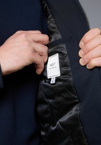 Mustas kohandatud jope sisemus, millel on sile satäänvooder, millel on silt "NEXT TAILORING" ja peidetud tasku.