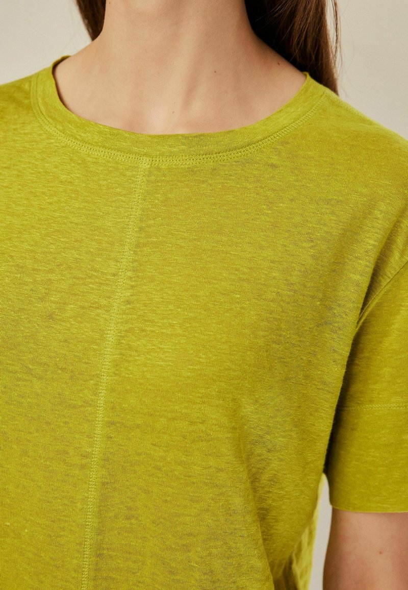 Femme portant un t-shirt vert citron à manches courtes avec un col rond et une couture verticale visible au centre devant.