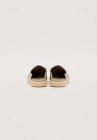 Bežās slip-on kurpes ar strukturētu auduma augšdaļu un jūtu virves zoli. Bez aizmugures atbalsta, redzams iekšējais oderējums un atvērts dizains.