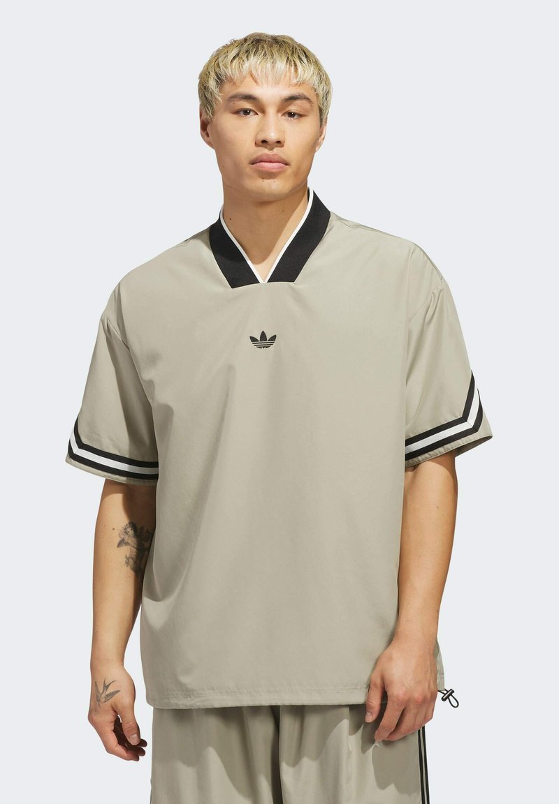 Svetlo béžová kratká košeľa s krátkym rukávom a čiernym výstrihom do V, so súvisle pruhmi na rukávoch a malým čiernym logom na hrudi. Hladká textúra.