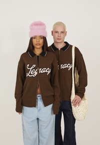 Des gilets zippés marron avec "Legacy" brodé en rose clair. Associés à un pantalon baggy en denim clair et foncé. Chapeau en peluche rose et sac en crochet.