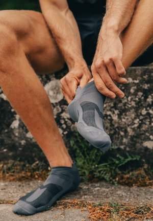 Graue Sportsocke mit strukturiertem Muster, passgenauer Form und verstärktem Fersenbereich. Wird von einer Person getragen, die im Sitzen ihre Position anpasst.