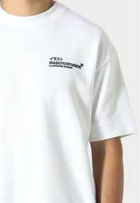 Biała bawełniana koszulka z krótkim rękawem, w luźnym kroju, z czarnym logo na piersi oraz tekstem w języku japońskim i angielskim.