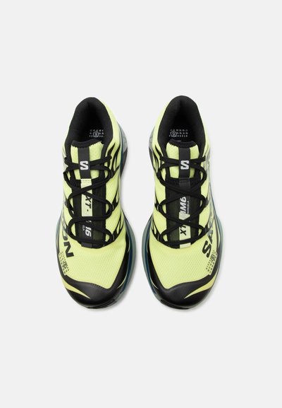 Φωτεινά κίτρινα και μαύρα αθλητικά παπούτσια με μαύρα κορδόνια και λεπτομέρειες της μάρκας, από πάνω σε λευκό φόντο.