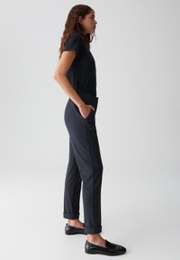 Woman in black t-shirt and dark cuffed trousers stands in profile with hands in pockets wearing black slip-on shoes against plain background.