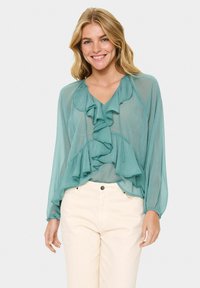 Teal, lightweight blouse with long sleeves and ruffled front design. Features a v-neckline and flows loosely over light-colored pants.