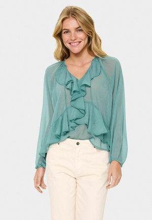 Teal, lightweight blouse with long sleeves and ruffled front design. Features a v-neckline and flows loosely over light-colored pants.