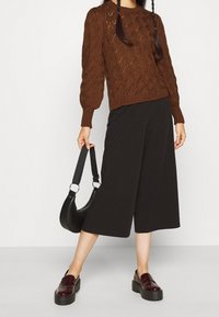 Osoba w brązowym swetrze z dzianiny, czarnych szerokich spodniach o długości 3/4, burgundowych butach na platformie i trzymająca czarną torebkę za ramiączko.