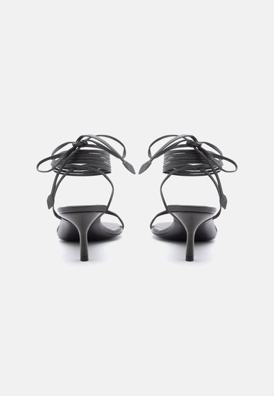 Μαύρα σανδάλια με λεπτά λουράκια και ψηλό τακούνι, με λεπτά λουράκια που τυλίγονται γύρω από τον αστράγαλο, εμφανιζόμενα από πίσω σε λευκό φόντο.