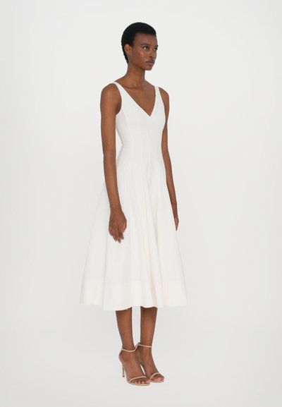 Witte mouwloze jurk met een V-hals, getailleerde lijfje, uitlopende rok en gladde stof. Gecombineerd met licht nude enkelsandalen met bandjes.