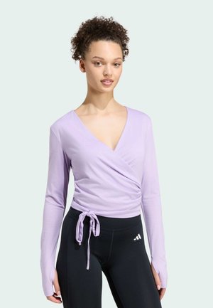 Kvinde med krøllet hår iført en lyse lilla viklet top med hul til tommelfingrene og sorte Adidas leggings mod en ensfarvet lys baggrund.