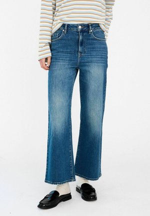 Osoba nosząca wyblakłe proste dżinsy w kolorze niebieskim, czarne mokasyny, białe skarpetki oraz koszulę w paski z długim rękawem w kolorach beżowym, niebieskim i białym.