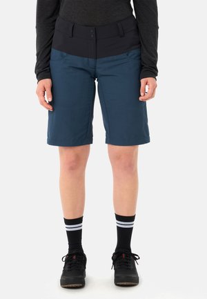 Person trägt dunkelblaue Shorts mittlerer Länge mit schwarzem Bund, schwarze Sportschuhe, schwarze Socken mit zwei weißen Streifen und ein langärmeliges Oberteil.
