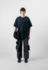 Camiseta negra de gran tamaño con diseño gráfico, combinada con pantalones cargo oscuros que tienen bolsillos y cremalleras. Modelo sosteniendo una chaqueta negra.
