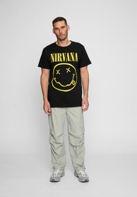 Mand, der står iført en sort Nirvana T-shirt med gul smiley-ansigt, lysegrå bukser og hvid-grå sneakers foran en ensfarvet baggrund.